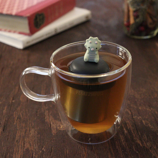 triceratops dinosaur animal tea infuser for loose leaf tea  with a fine mesh stainless steel tea infuser that reads FLOAT2INFUSE on a black base floating in a glass tea mug with books and loose leaf tea in the background