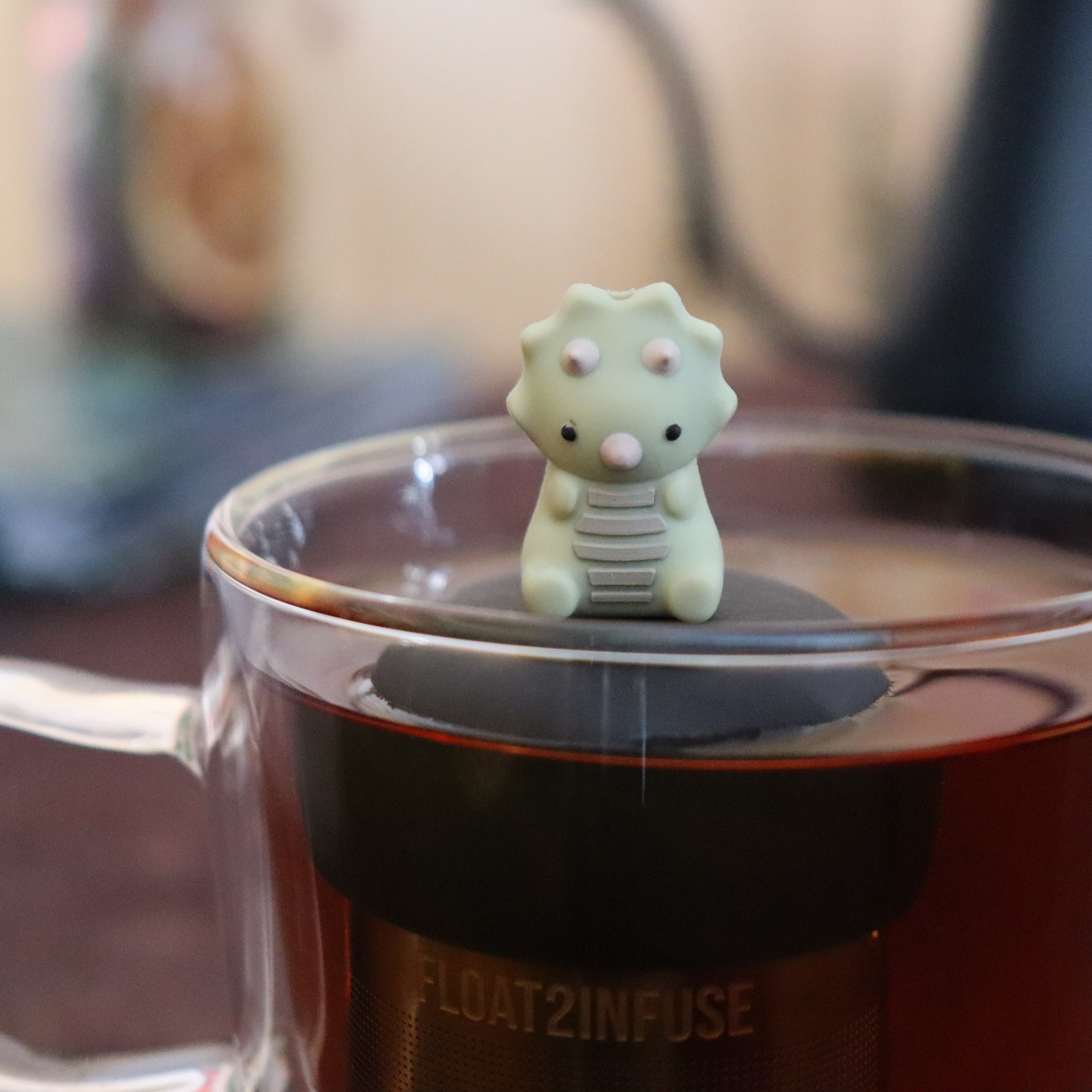 sage green triceratops dinosaur animal tea infuser for loose leaf tea  with a fine mesh stainless steel tea infuser that reads FLOAT2INFUSE on a black base floating in a glass tea mug with tea kettle in the background