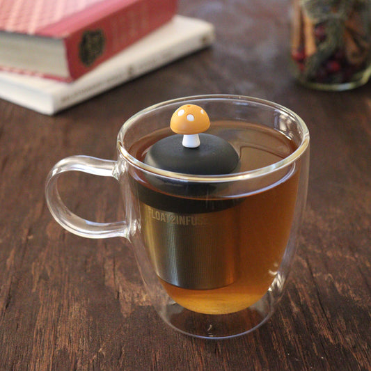 yellow mushroom tea infuser for loose leaf tea  with a fine mesh stainless steel tea infuser that reads FLOAT2INFUSE on a black base floating in a glass tea mug with books and loose leaf tea in the background