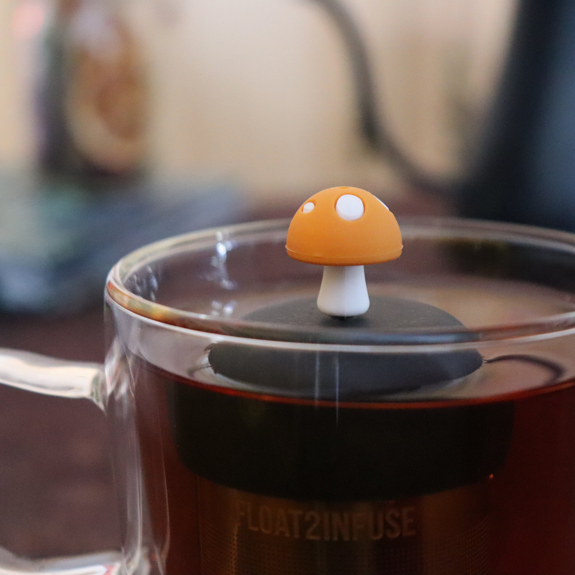 yellow mushroom tea infuser for loose leaf tea  with a fine mesh stainless steel strainer that reads FLOAT2INFUSE on a black base floating in a glass tea mug with tea kettle in the background