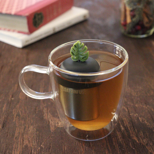 army green monstera leaf tea infuser for loose leaf tea on a black base with a fine mesh stainless steel tea infuser that reads FLOAT2INFUSE floating in a glass tea mug with books and loose leaf tea in the background