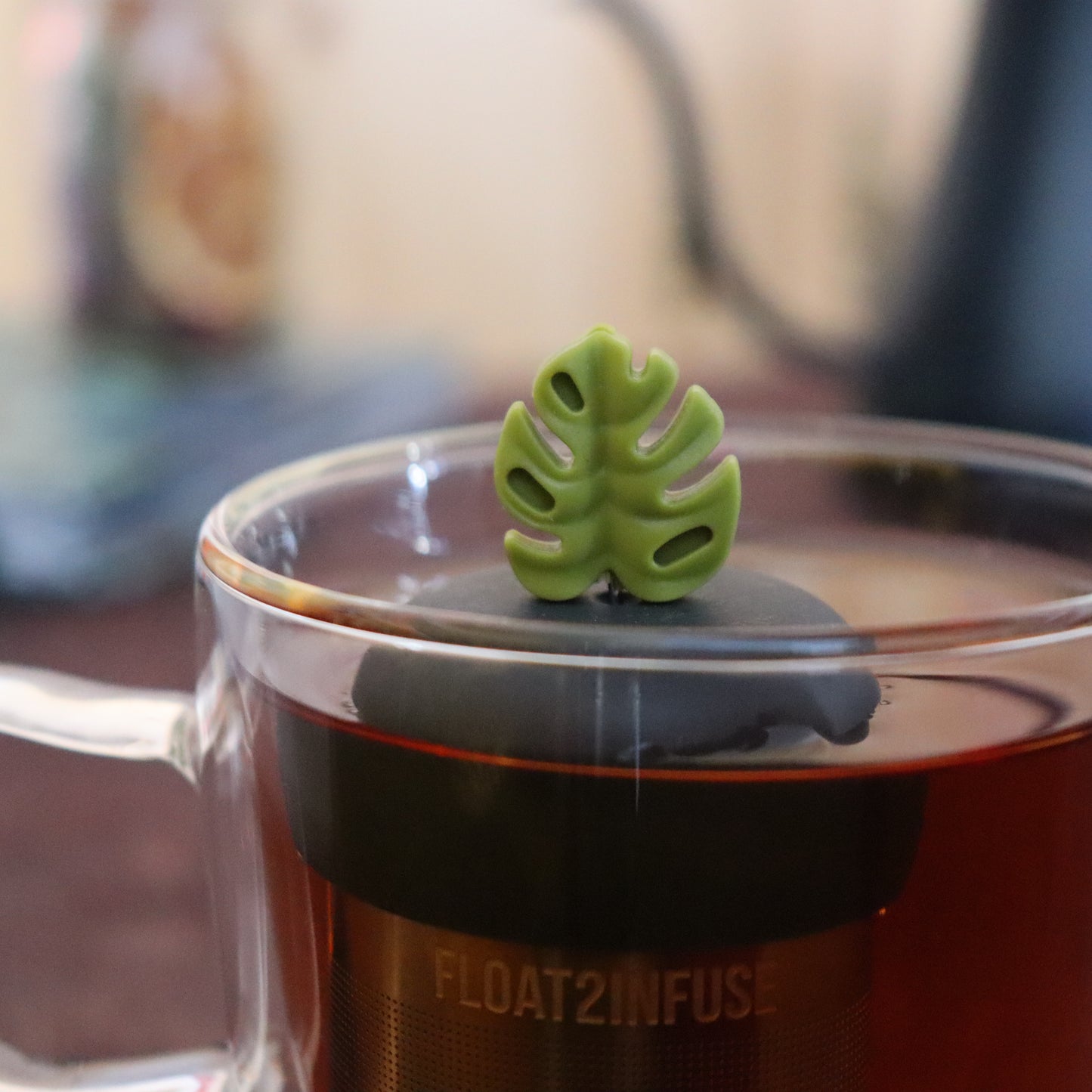 army green monstera leaf tea infuser for loose leaf tea with a fine mesh stainless steel strainer that reads FLOAT2INFUSE on a black base floating in a glass tea mug with tea kettle in the background