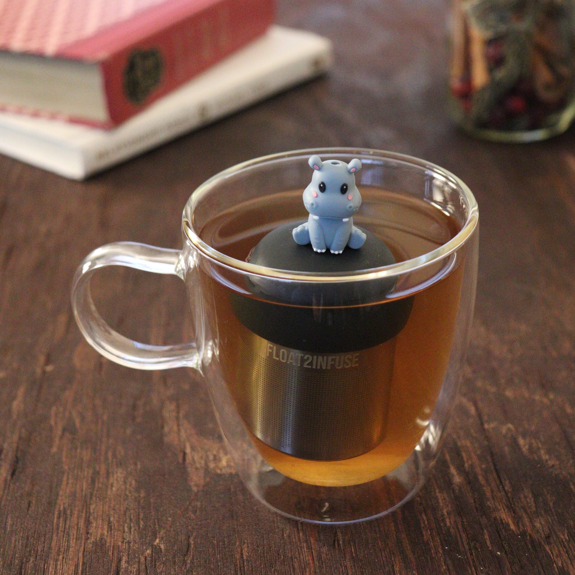 blushing grey baby hippo animal tea infuser for loose leaf tea  with a fine mesh stainless steel tea infuser that reads FLOAT2INFUSE on a black base floating in a glass tea mug with books and loose leaf tea in the background