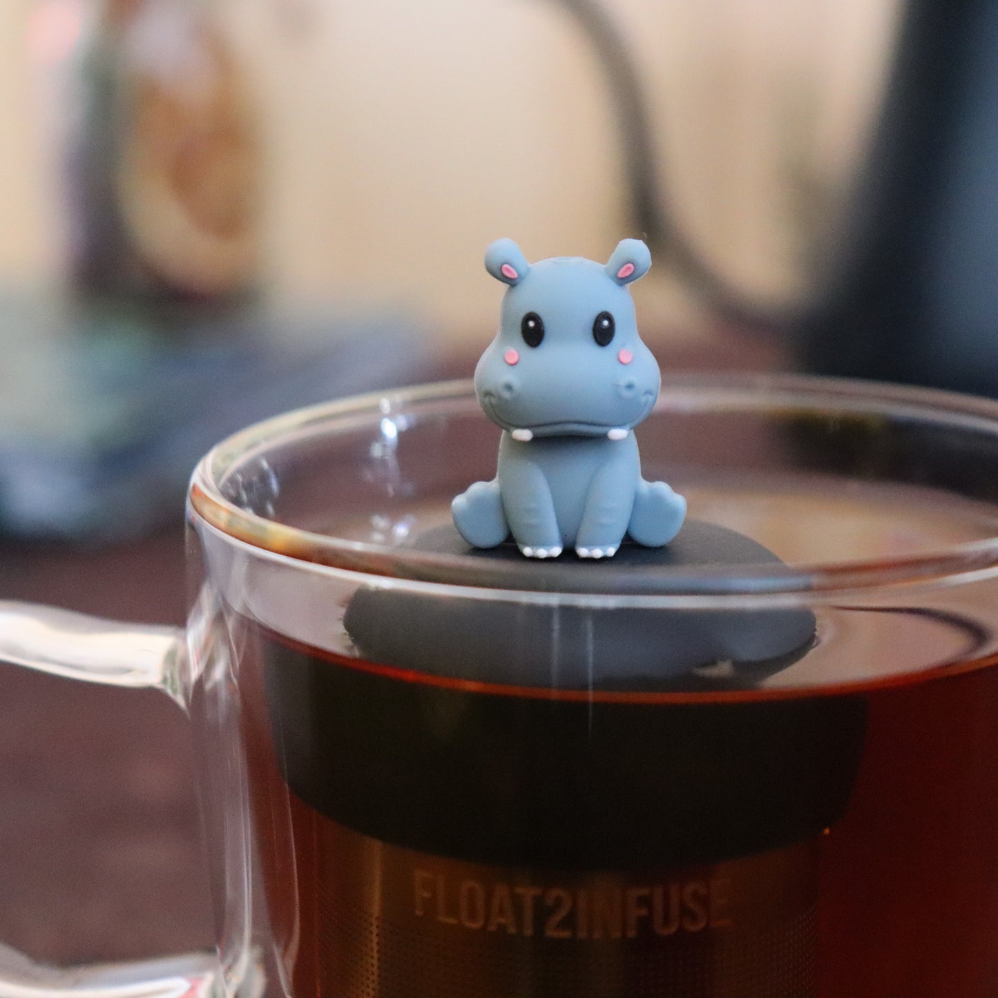 grey smiling baby hippo animal tea infuser for loose leaf tea  with a fine mesh stainless steel tea infuser that reads FLOAT2INFUSE on a black base floating in a glass tea mug with tea kettle in the background
