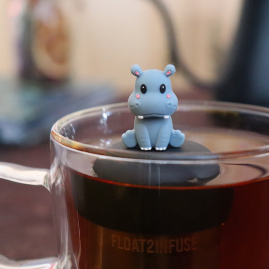 grey smiling baby hippo animal tea infuser for loose leaf tea  with a fine mesh stainless steel tea infuser that reads FLOAT2INFUSE on a black base floating in a glass tea mug with tea kettle in the background