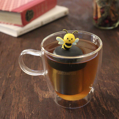 black and yellow bee animal tea infuser for loose leaf tea  with a fine mesh stainless steel tea infuser that reads FLOAT2INFUSE on a black base floating in a glass tea mug with books and loose leaf tea in the background