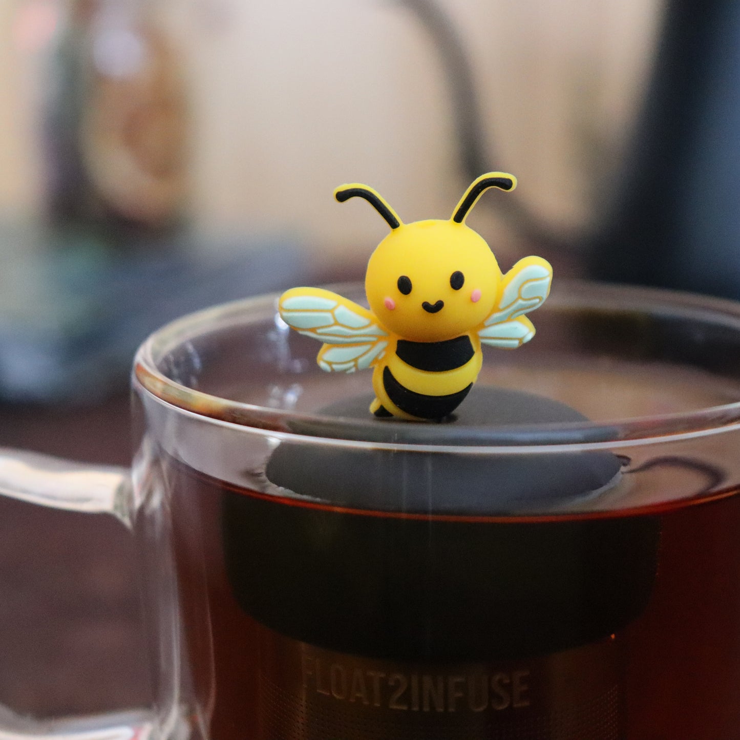 yellow and black bee animal tea infuser for loose leaf tea with a fine mesh stainless steel tea infuser that reads FLOAT2INFUSE  on a black base floating in a glass tea mug with tea kettle in the background