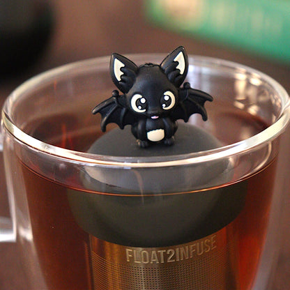 Black bat with outstretched wings tea infuser for loose leaf tea with a fine mesh stainless steel tea strainer 
that reads FLOAT2INFUSE on a black base with a black cauldron tea mug and books in the background