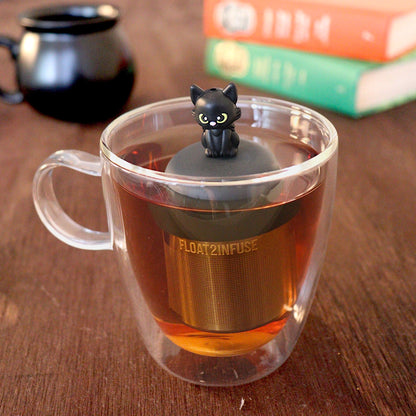Black cat tea infuser for loose leaf tea with a fine mesh stainless steel tea strainer that reads FLOAT2INFUSE on a black base with a black cauldron shaped tea mug and books in the background