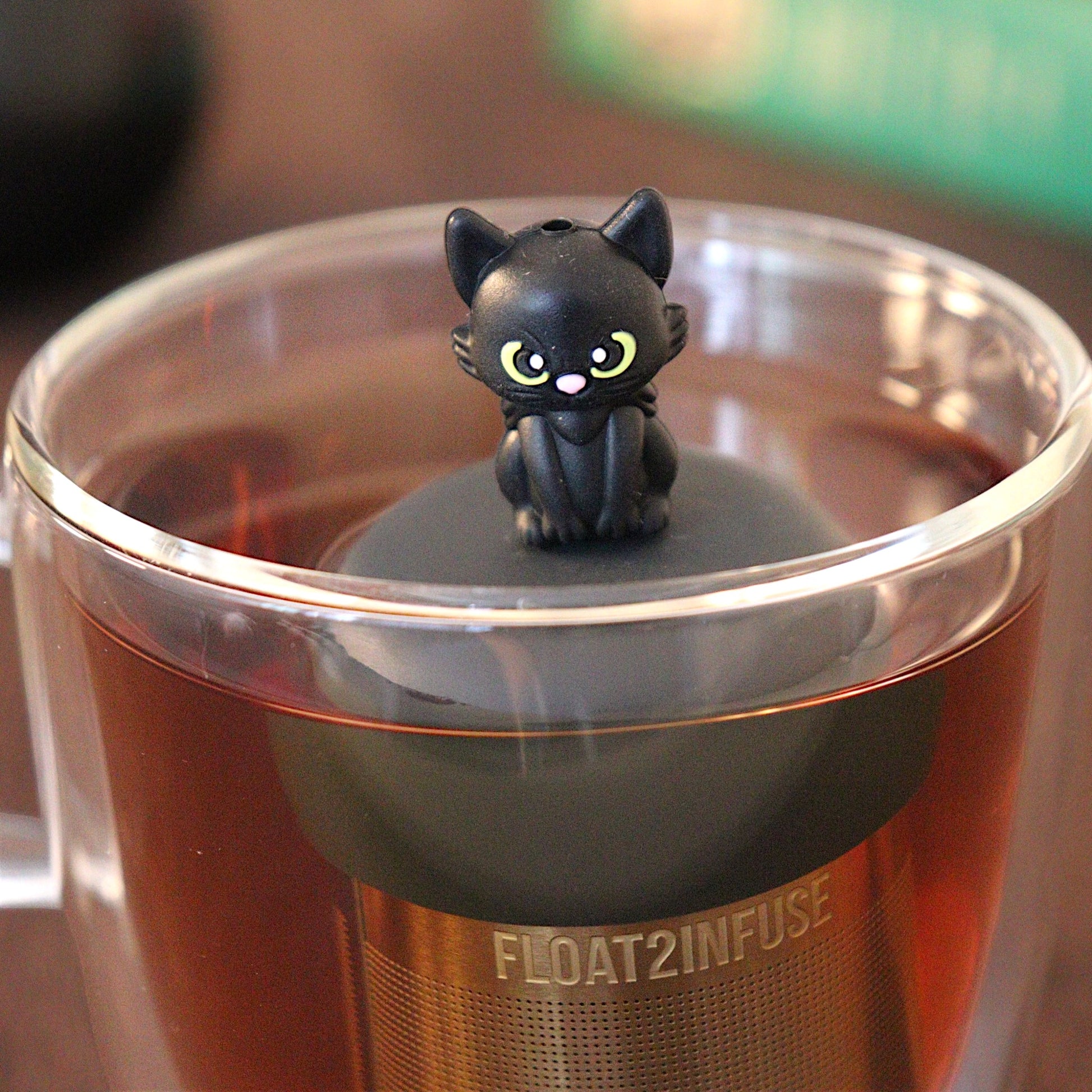 Black cat tea infuser for loose leaf tea with a fine mesh stainless steel tea strainer that reads FLOAT2INFUSE on a black base with a black cauldron shaped tea mug and books in the background