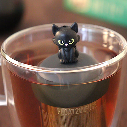 Black cat tea infuser for loose leaf tea with a fine mesh stainless steel tea strainer that reads FLOAT2INFUSE on a black base with a black cauldron shaped tea mug and books in the background
