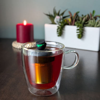 backside of black cauldron with green goo tea infuser for loose leaf tea  with a fine mesh stainless steel strainer that reads FLOAT2INFUSE on a black base floating in a glass tea with plants and a candle in the background mug