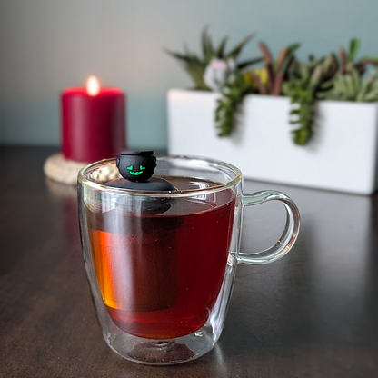 smiling black cauldron tea infuser for loose leaf tea  with a fine mesh stainless steel tea infuser that reads FLOAT2INFUSE on a black base floating in a glass tea mug with plants and a candle in the background