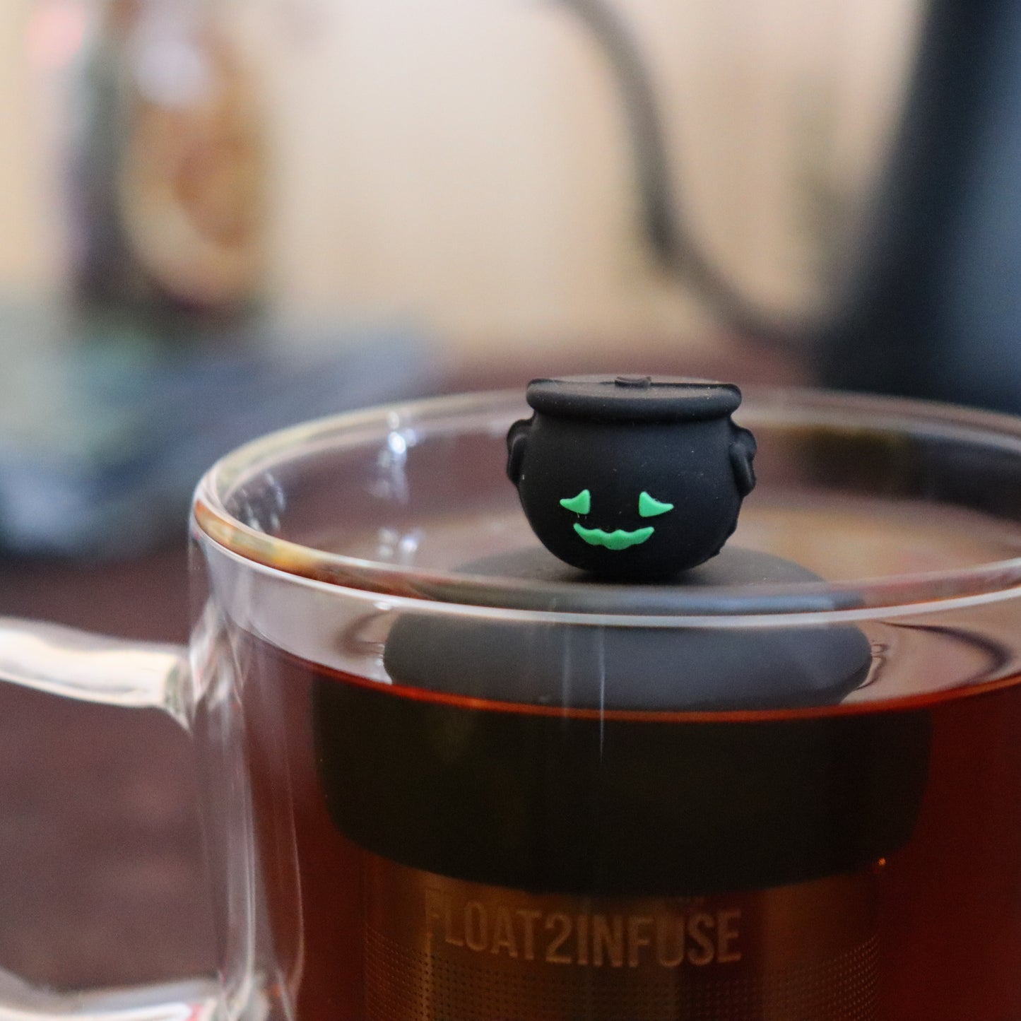 black smiling cauldron tea infuser for loose leaf tea e with a fine mesh stainless steel strainer that reads FLOAT2INFUSE on a black bas floating in a glass tea mug with tea kettle in the background
