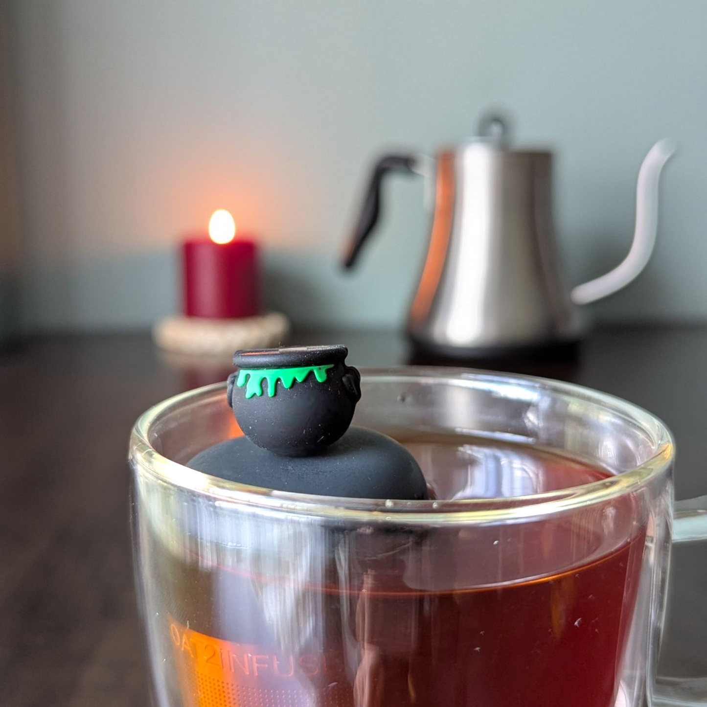 black cauldron with green goo tea infuser for loose leaf tea  with a fine mesh stainless steel tea infuser that reads FLOAT2INFUSE on a black base floating in a glass tea mug with a candle and tea kettle in the background