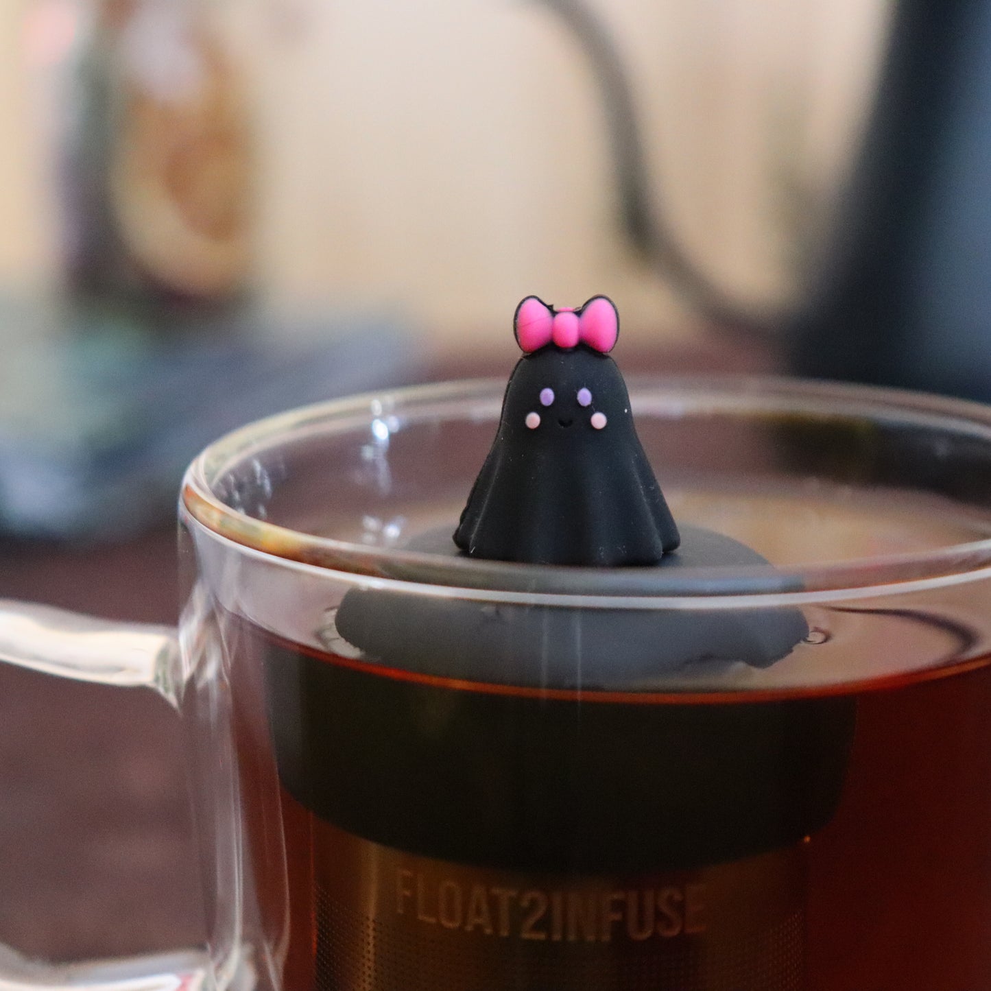 blushing black ghost with pink bow tea infuser for loose leaf tea  with a fine mesh stainless steel strainer that reads FLOAT2INFUSE on a black base floating in a glass tea mug with tea kettle in the background