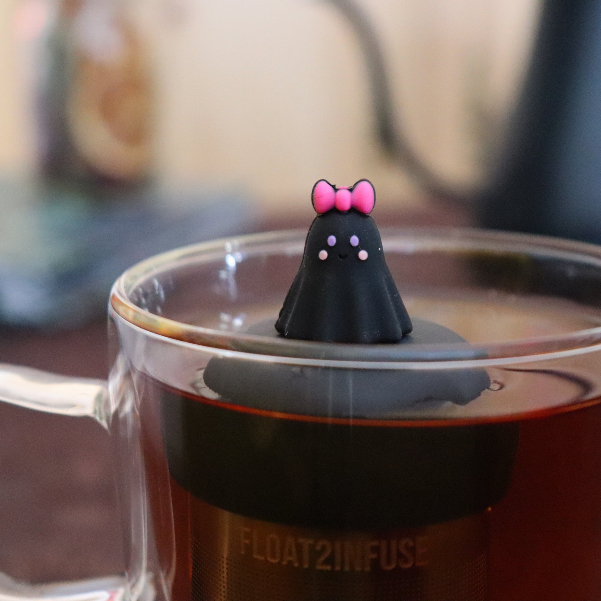blushing black ghost with pink bow tea infuser for loose leaf tea  with a fine mesh stainless steel strainer that reads FLOAT2INFUSE on a black base floating in a glass tea mug with tea kettle in the background