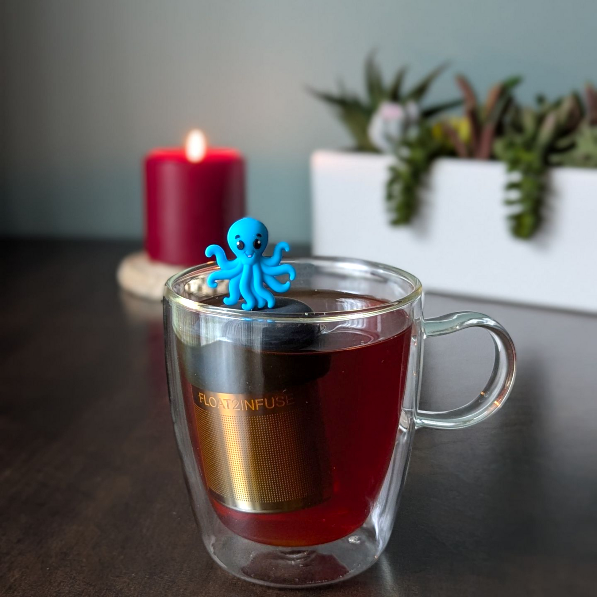 blue octopus animal tea infuser for loose leaf tea  with a fine mesh stainless steel tea infuser that reads FLOAT2INFUSE on a black base floating in a glass tea mug with a plant and a candle in the background
