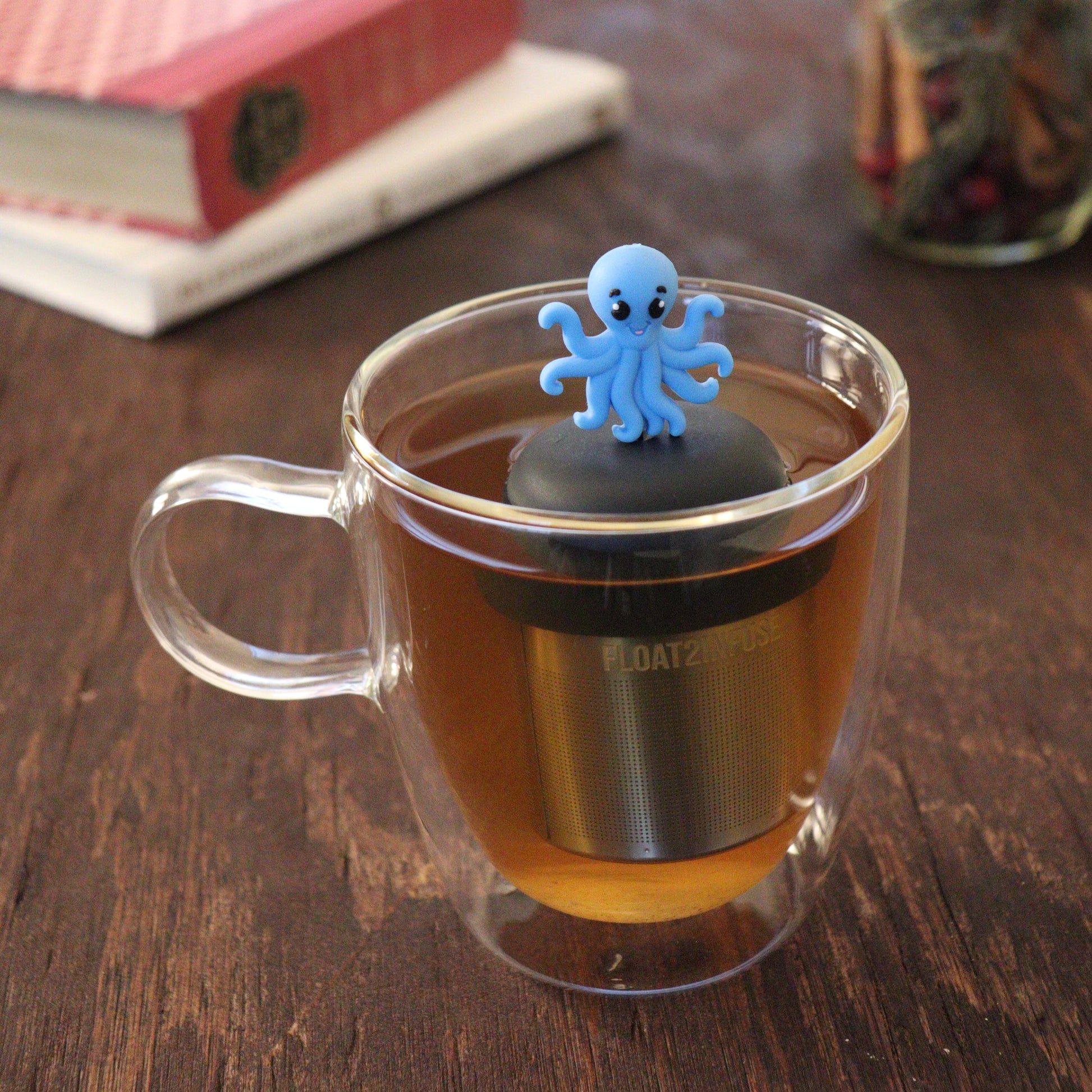 blue octopus animal tea infuser for loose leaf tea  with a fine mesh stainless steel tea infuser that reads FLOAT2INFUSE on a black base floating in a glass tea mug with books and loose leaf tea in the background
