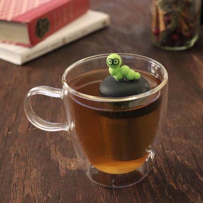 green bookworm wearing glasses animal tea infuser for loose leaf tea  with a fine mesh stainless steel tea infuser that reads FLOAT2INFUSE on a black base floating in a glass tea mug with books and loose leaf tea in the background
