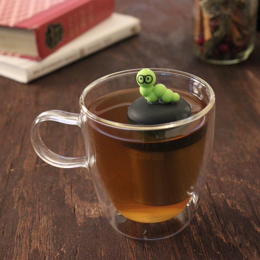green bookworm wearing glasses animal tea infuser for loose leaf tea  with a fine mesh stainless steel tea infuser that reads FLOAT2INFUSE on a black base floating in a glass tea mug with books and loose leaf tea in the background
