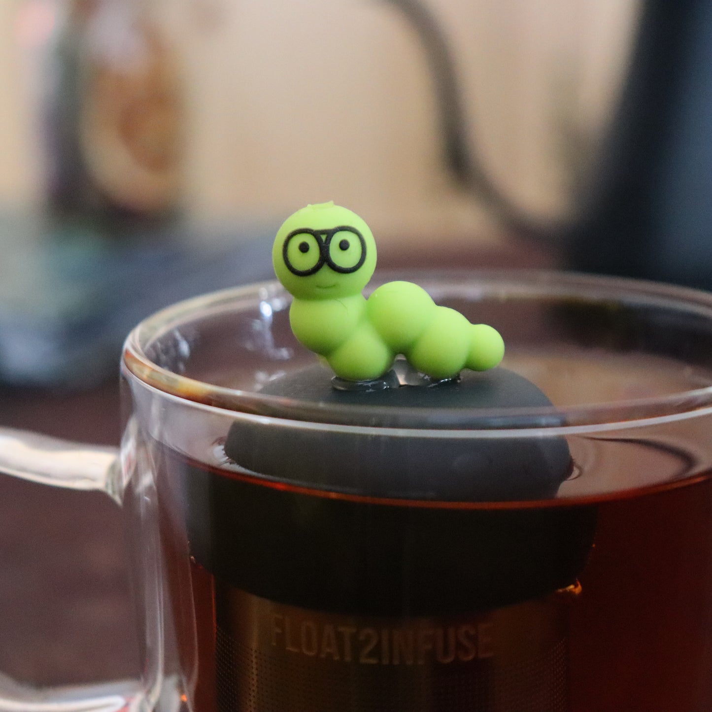 green bookworm with glasses animal tea infuser for loose leaf tea  with a fine mesh stainless steel tea infuser that reads FLOAT2INFUSE on a black base floating in a glass tea mug with tea kettle in the background