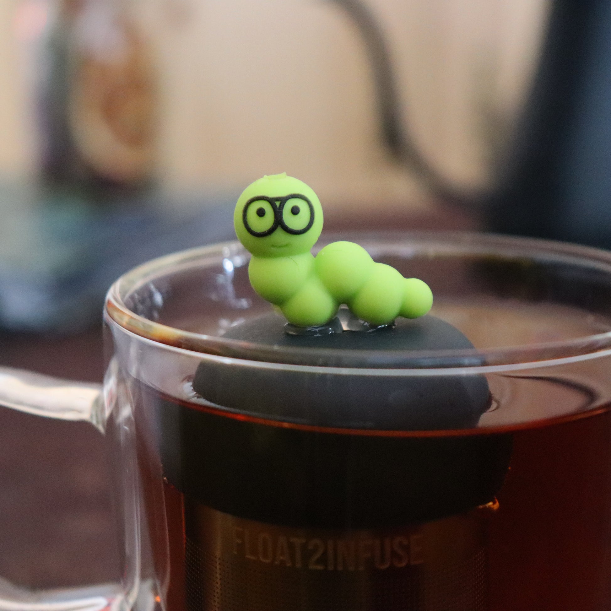 green bookworm with glasses animal tea infuser for loose leaf tea  with a fine mesh stainless steel tea infuser that reads FLOAT2INFUSE on a black base floating in a glass tea mug with tea kettle in the background