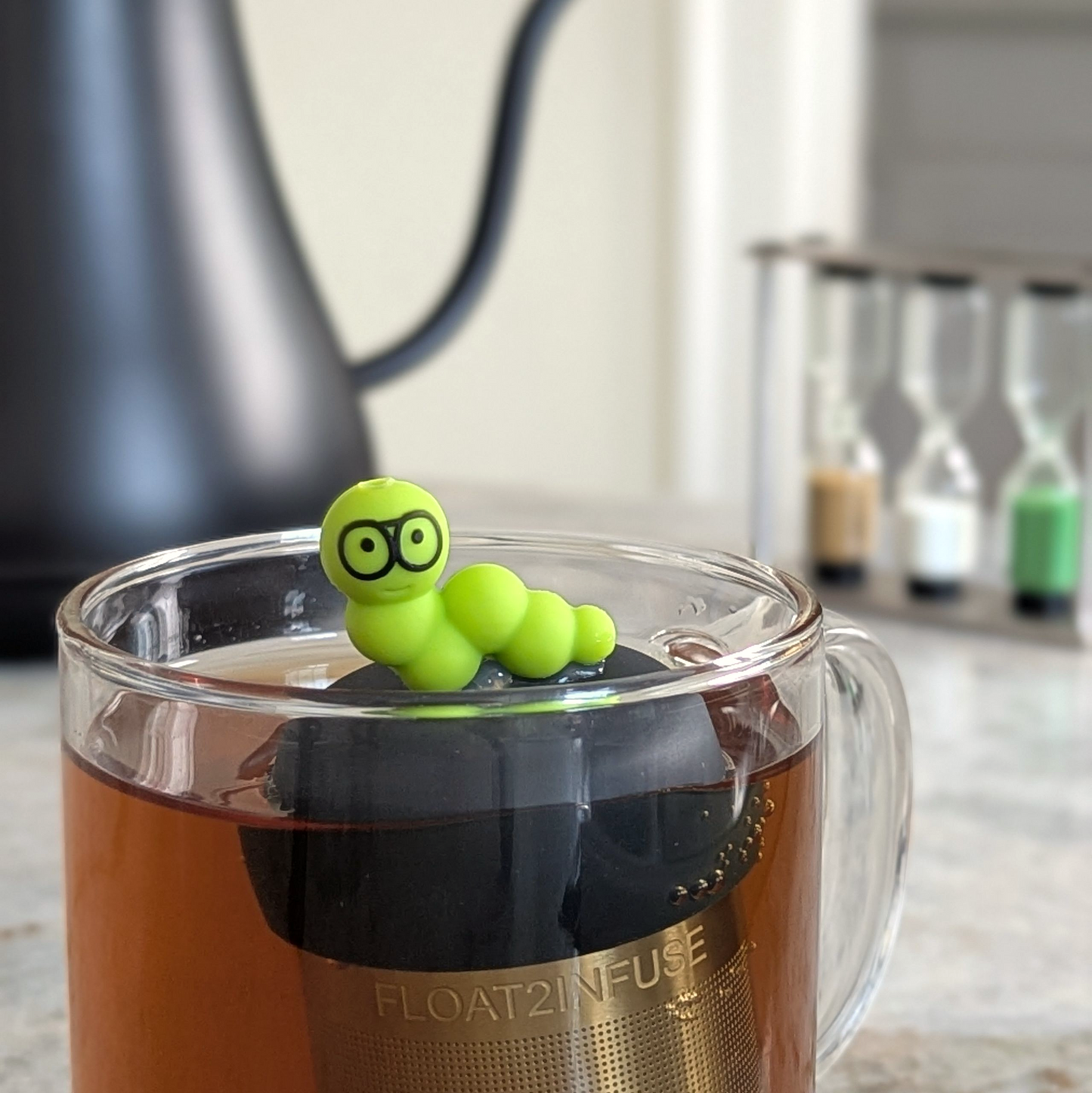 green bookworm tea infuser for loose leaf tea  with a fine mesh stainless steel strainer that reads FLOAT2INFUSE on a black base floating in a glass tea mug with tea kettle in the background