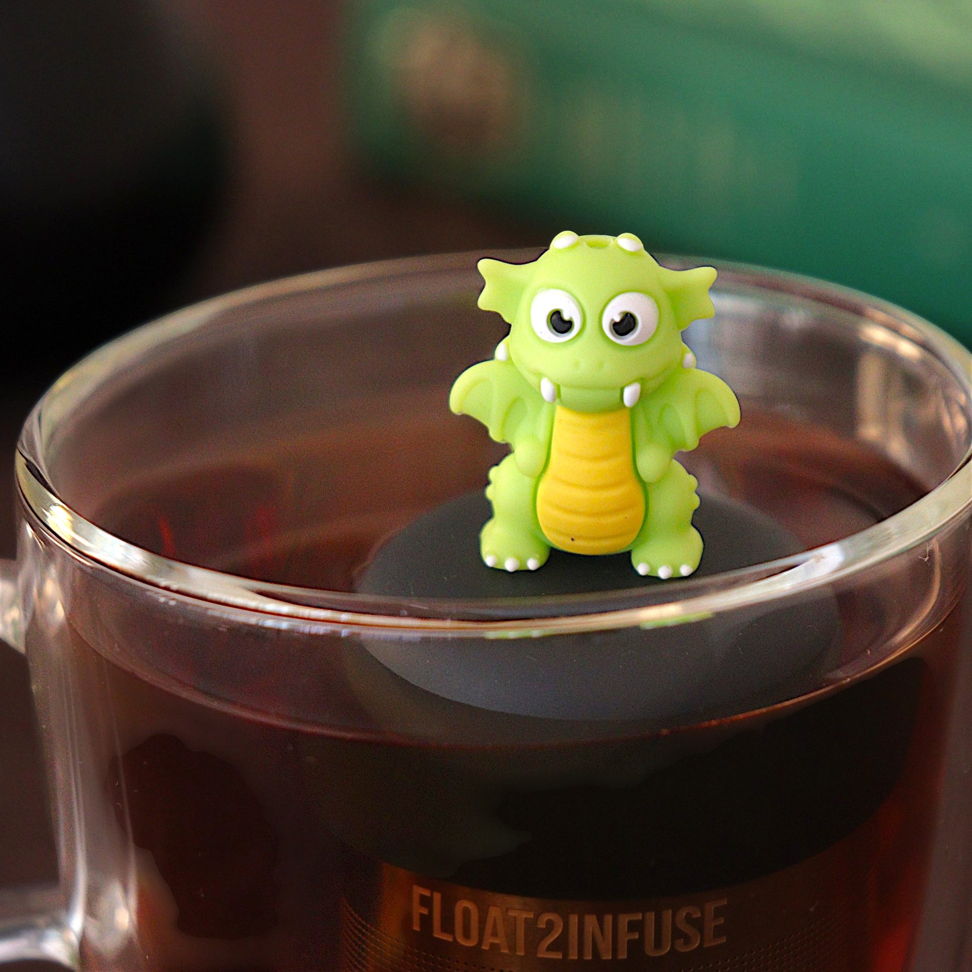 bright green dragon tea infuser for loose leaf tea  with a fine mesh stainless steel tea infuser that reads FLOAT2INFUSE on a black base floating in a glass tea mug with books in the background