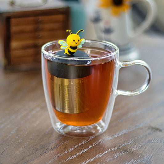 black and yellow bee animal tea infuser for loose leaf tea  with a fine mesh stainless steel tea infuser that reads FLOAT2INFUSE on a black base floating in a glass tea mug with a water pitcher tea in the background