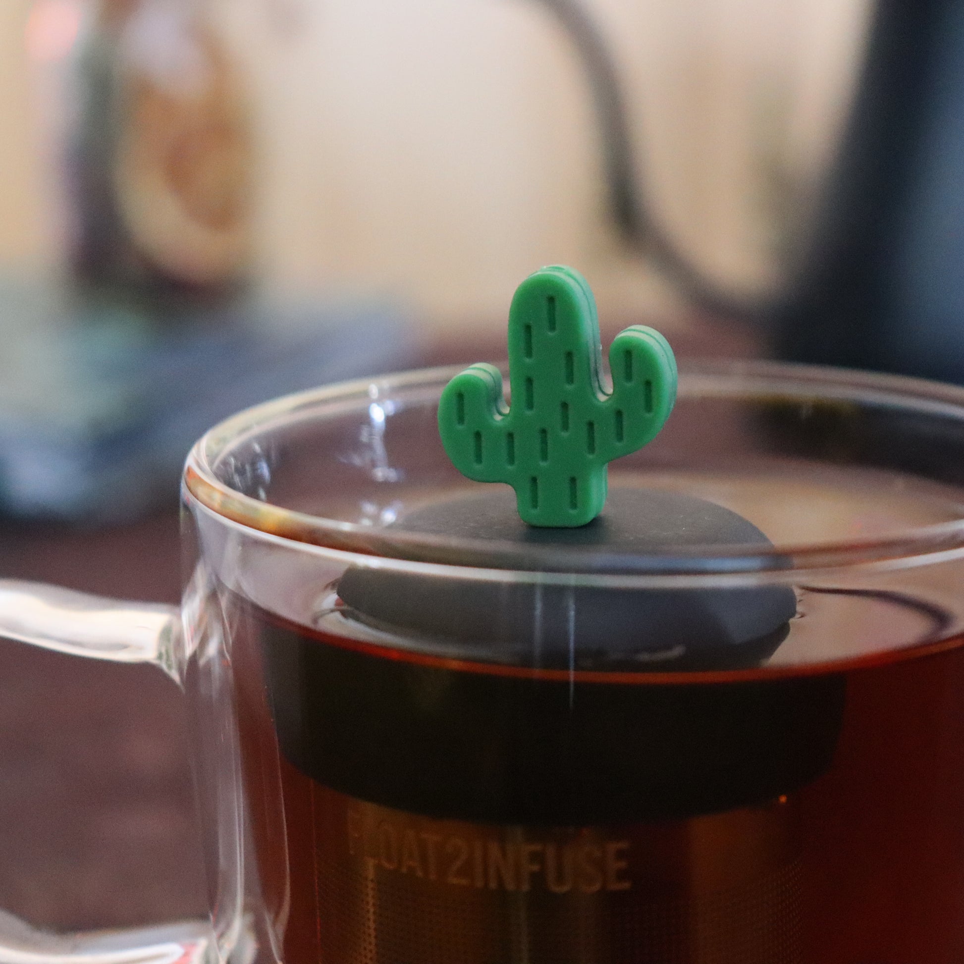 green cactus tea infuser for loose leaf tea  with a fine mesh stainless steel strainer that reads FLOAT2INFUSE on a black base floating in a glass tea mug with tea kettle in the background