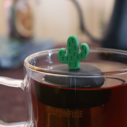 green cactus tea infuser for loose leaf tea  with a fine mesh stainless steel strainer that reads FLOAT2INFUSE on a black base floating in a glass tea mug with tea kettle in the background