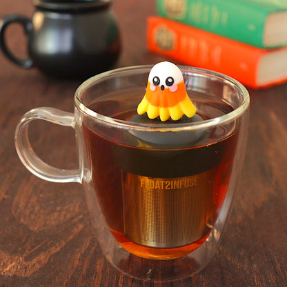 candy corn ghost tea infuser for loose leaf tea  with a fine mesh stainless steel tea infuser that reads FLOAT2INFUSE on a black base floating in a glass tea mug with books in the background 