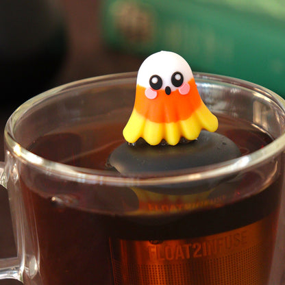 candy corn ghost tea infuser for loose leaf tea  with a fine mesh stainless steel tea infuser that reads FLOAT2INFUSE on a black base floating in a glass tea mug with a book in the background 