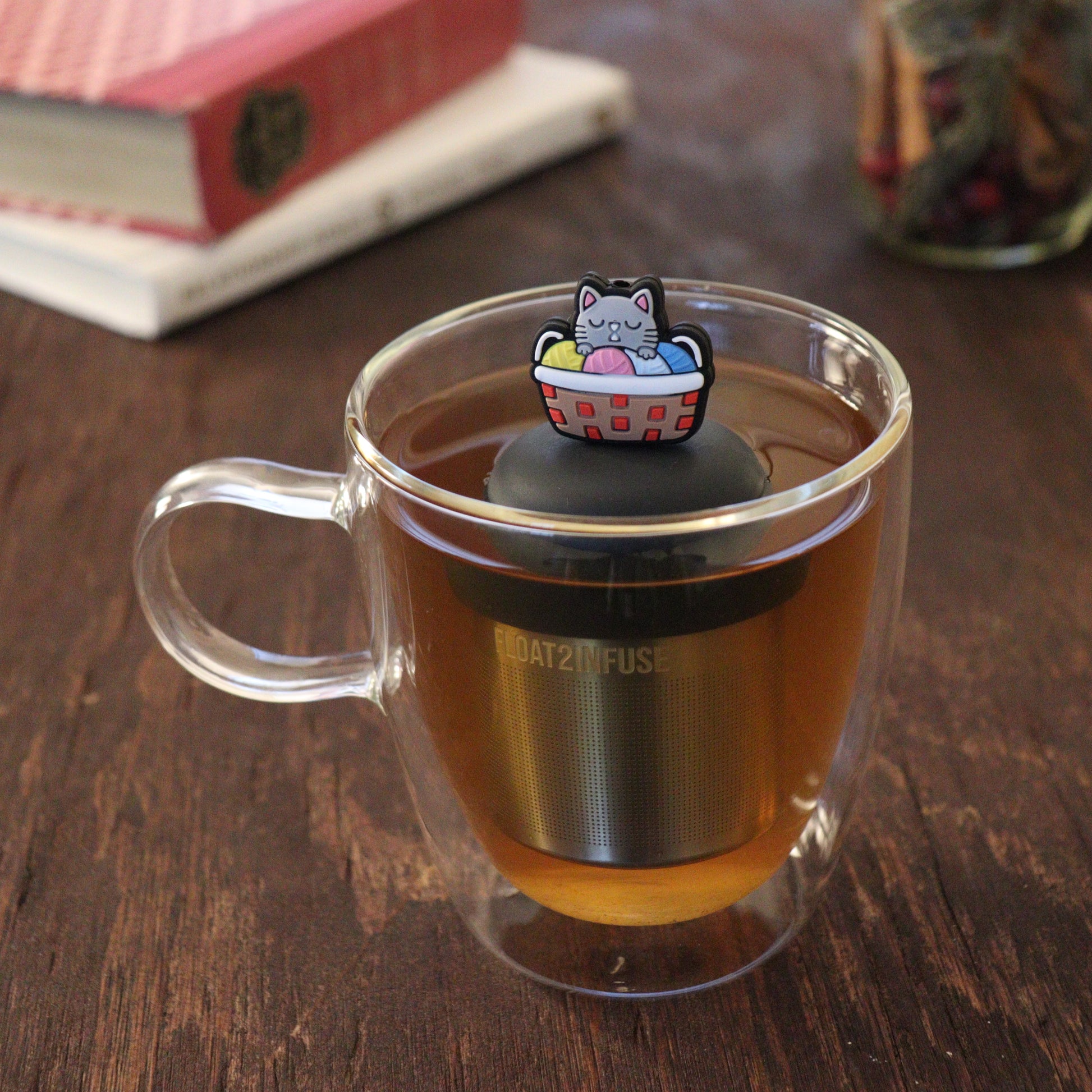 grey cat sitting in a multi colored knitting basket animal tea infuser for loose leaf tea  with a fine mesh stainless steel tea infuser that reads FLOAT2INFUSE on a black base floating in a glass tea mug with books and loose leaf tea in the background