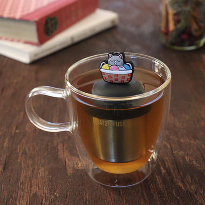 grey cat sitting in a multi colored knitting basket animal tea infuser for loose leaf tea  with a fine mesh stainless steel tea infuser that reads FLOAT2INFUSE on a black base floating in a glass tea mug with books and loose leaf tea in the background