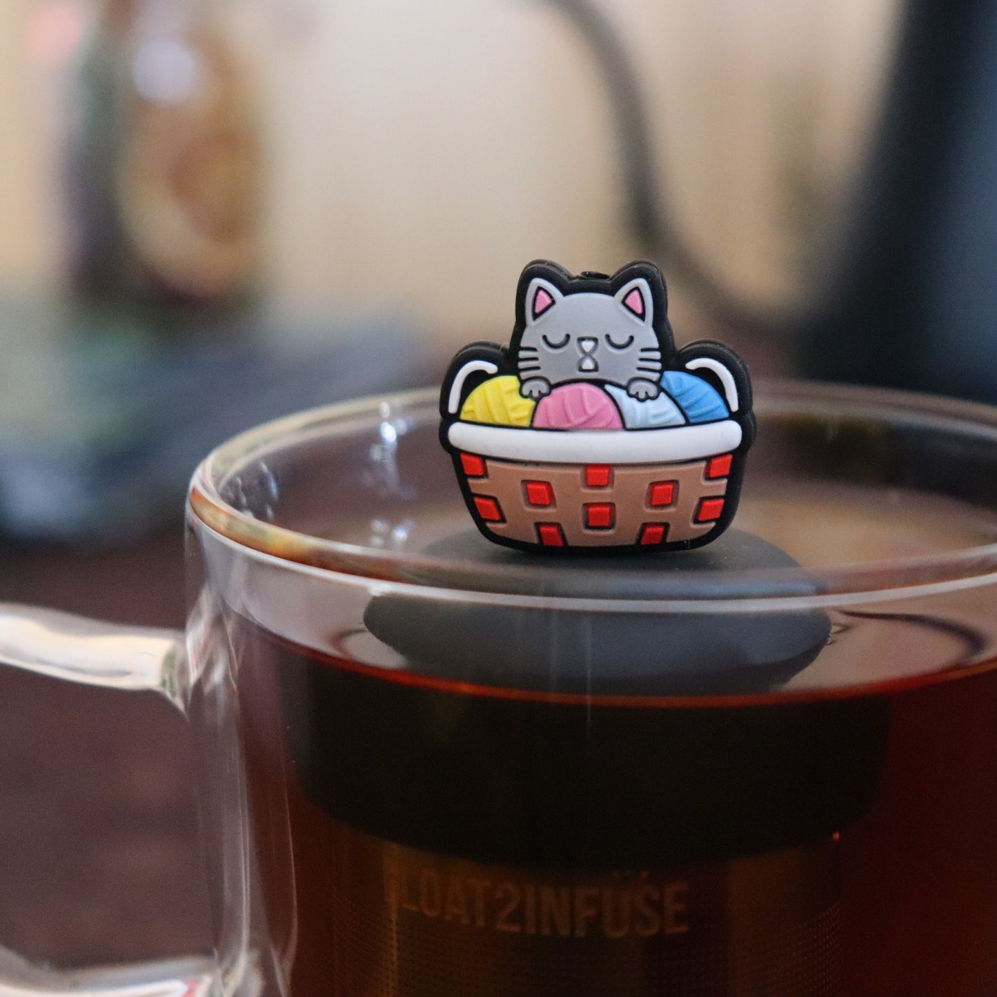 grey cat in colorful knitting basket animal  tea infuser for Loose leaf tea with a fine mesh stainless steel Tea steeper that reads FLOAT2INFUSE on black base floating in a Glass tea mug with tea kettle in the background