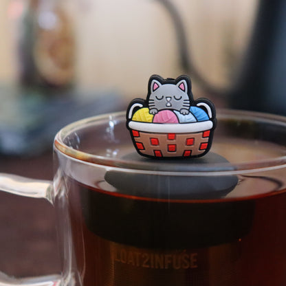 grey cat in colorful knitting basket animal  tea infuser for Loose leaf tea with a fine mesh stainless steel Tea steeper that reads FLOAT2INFUSE on black base floating in a Glass tea mug with tea kettle in the background