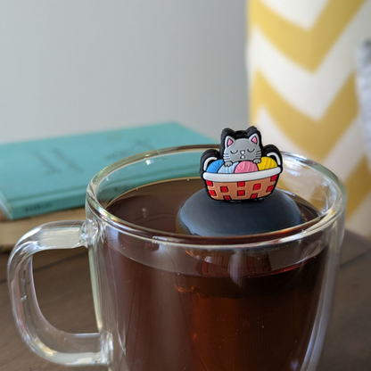 grey cat sitting in a multi colored knitting basket animal tea infuser for loose leaf tea  with a fine mesh stainless steel tea infuser that reads FLOAT2INFUSE on a black base floating in a glass tea mug with books in the background