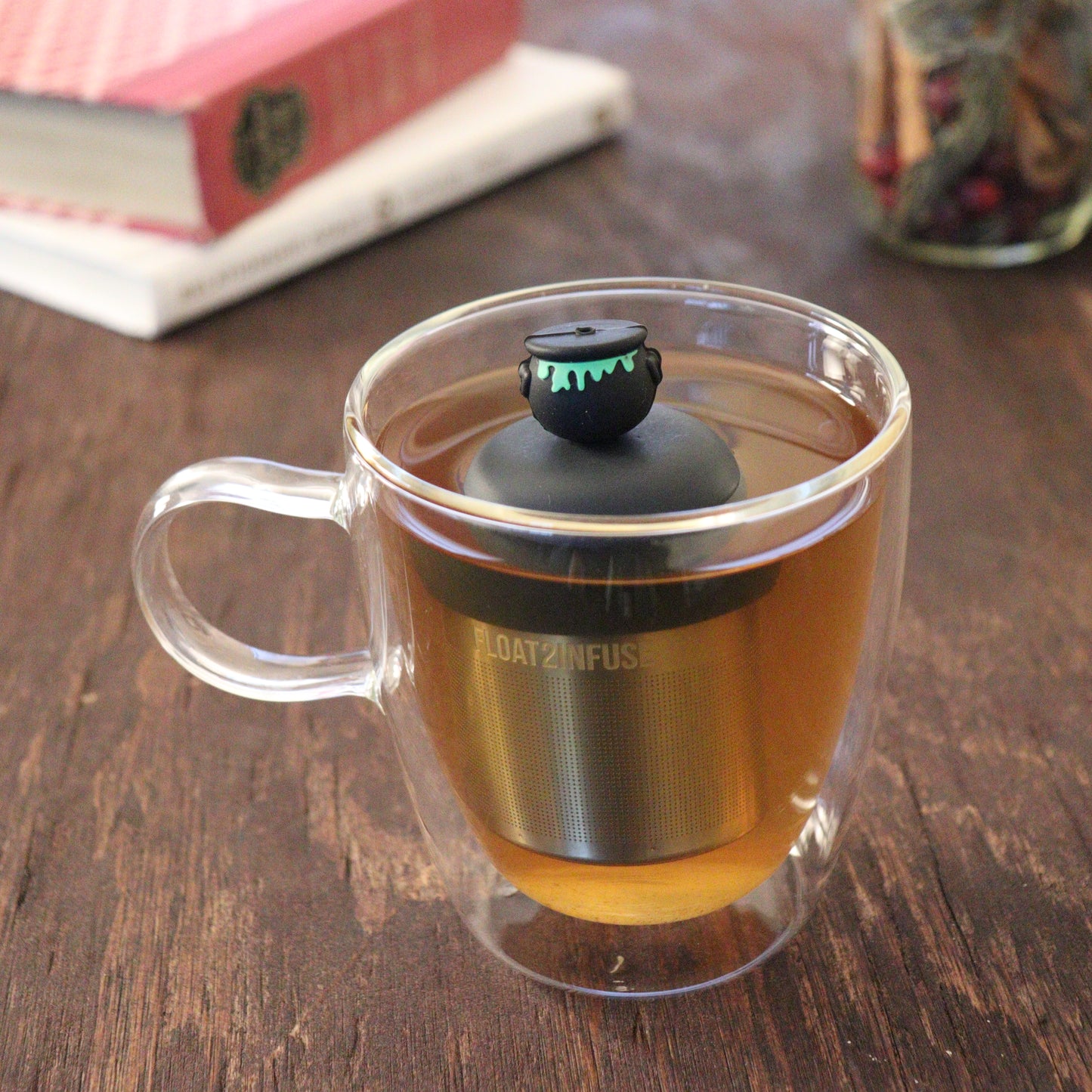 black cauldron with green goo tea infuser for loose leaf tea  with a fine mesh stainless steel tea infuser that reads FLOAT2INFUSE on a black base floating in a glass tea mug with books and loose leaf tea in the background