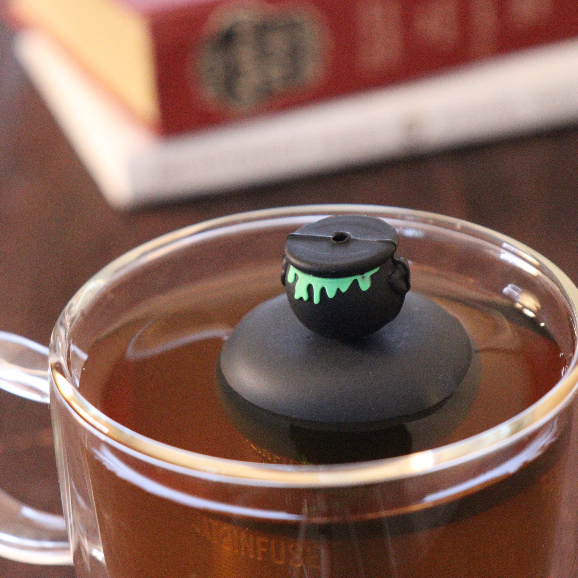 backside of black cauldron with green goo tea infuser for loose leaf tea  with a fine mesh stainless steel strainer that reads FLOAT2INFUSE on a black base floating in a glass tea mug with books in the background