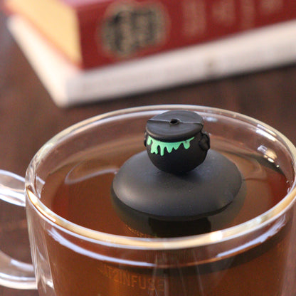backside of black cauldron with green goo tea infuser for loose leaf tea  with a fine mesh stainless steel strainer that reads FLOAT2INFUSE on a black base floating in a glass tea mug with books in the background