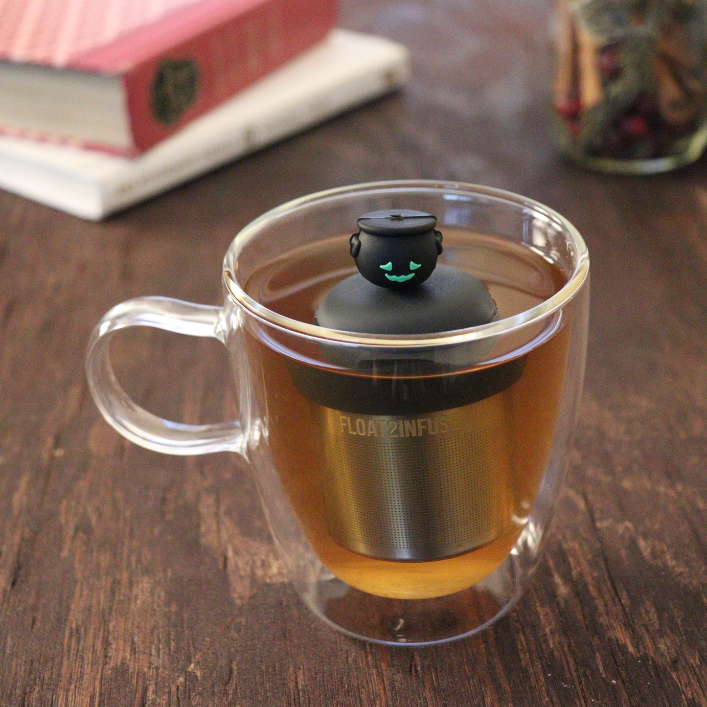 smiling black cauldron tea infuser for loose leaf tea  with a fine mesh stainless steel tea infuser that reads FLOAT2INFUSE on a black base floating in a glass tea mug with books and loose leaf tea in the background
