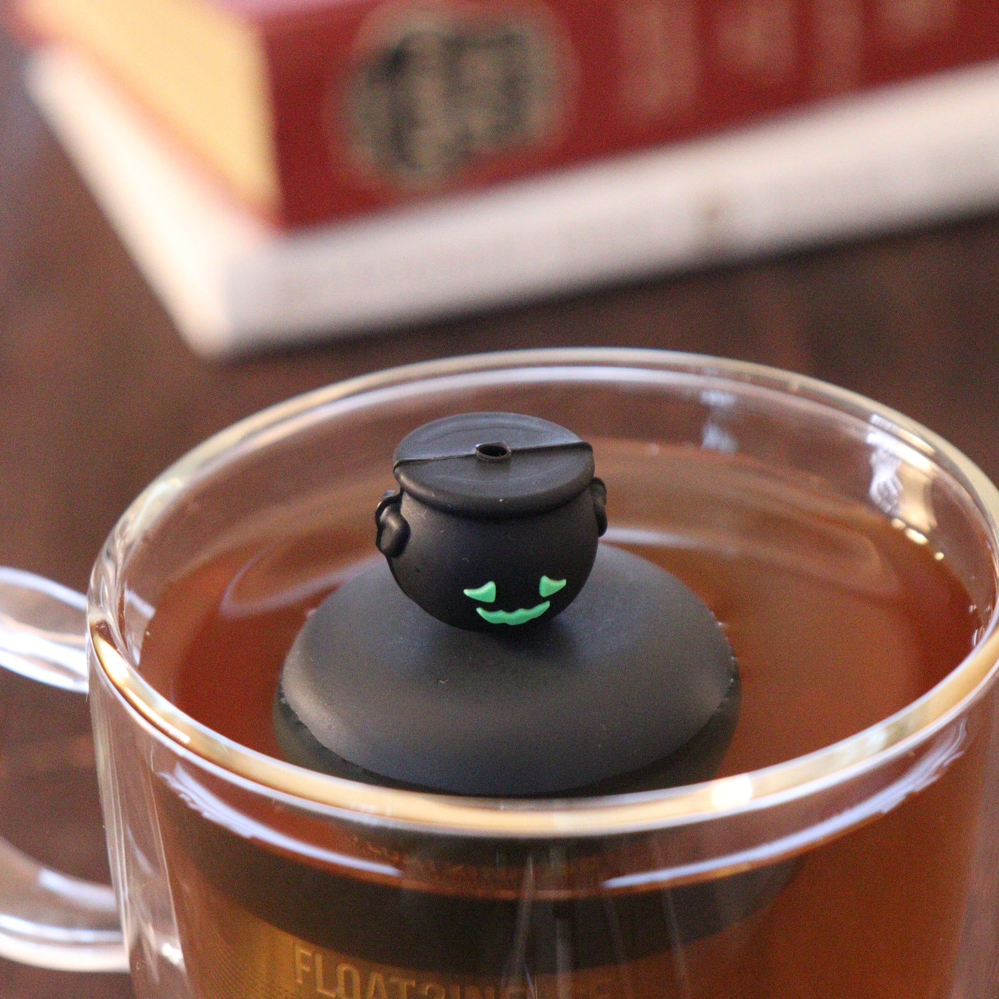 frontside of smiling black cauldron tea infuser for loose leaf tea  with a fine mesh stainless steel strainer that reads FLOAT2INFUSE on a black base floating in a glass tea mug with books in the background