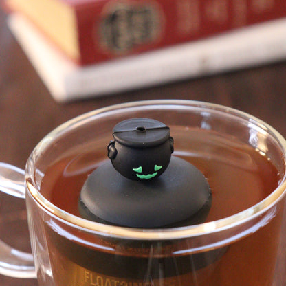 frontside of smiling black cauldron tea infuser for loose leaf tea  with a fine mesh stainless steel strainer that reads FLOAT2INFUSE on a black base floating in a glass tea mug with books in the background