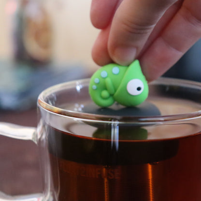green chameleon animal tea infuser for loose leaf tea  with a fine mesh stainless steel tea infuser that reads FLOAT2INFUSE on a black base floating in a glass tea mug with tea kettle in the background