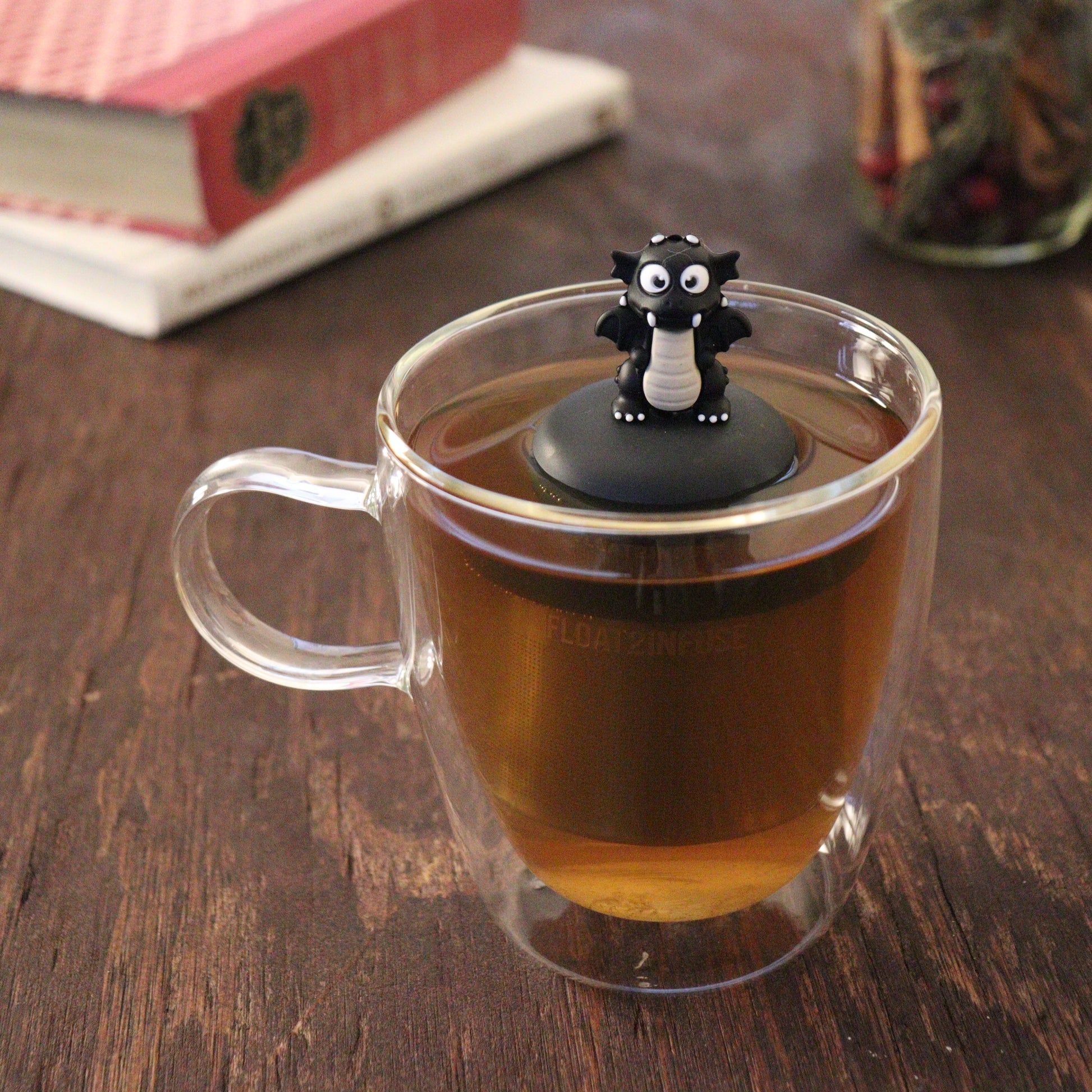 black dragon animal tea infuser for loose leaf tea  with a fine mesh stainless steel tea infuser that reads FLOAT2INFUSE on a black base floating in a glass tea mug with books and loose leaf tea in the background