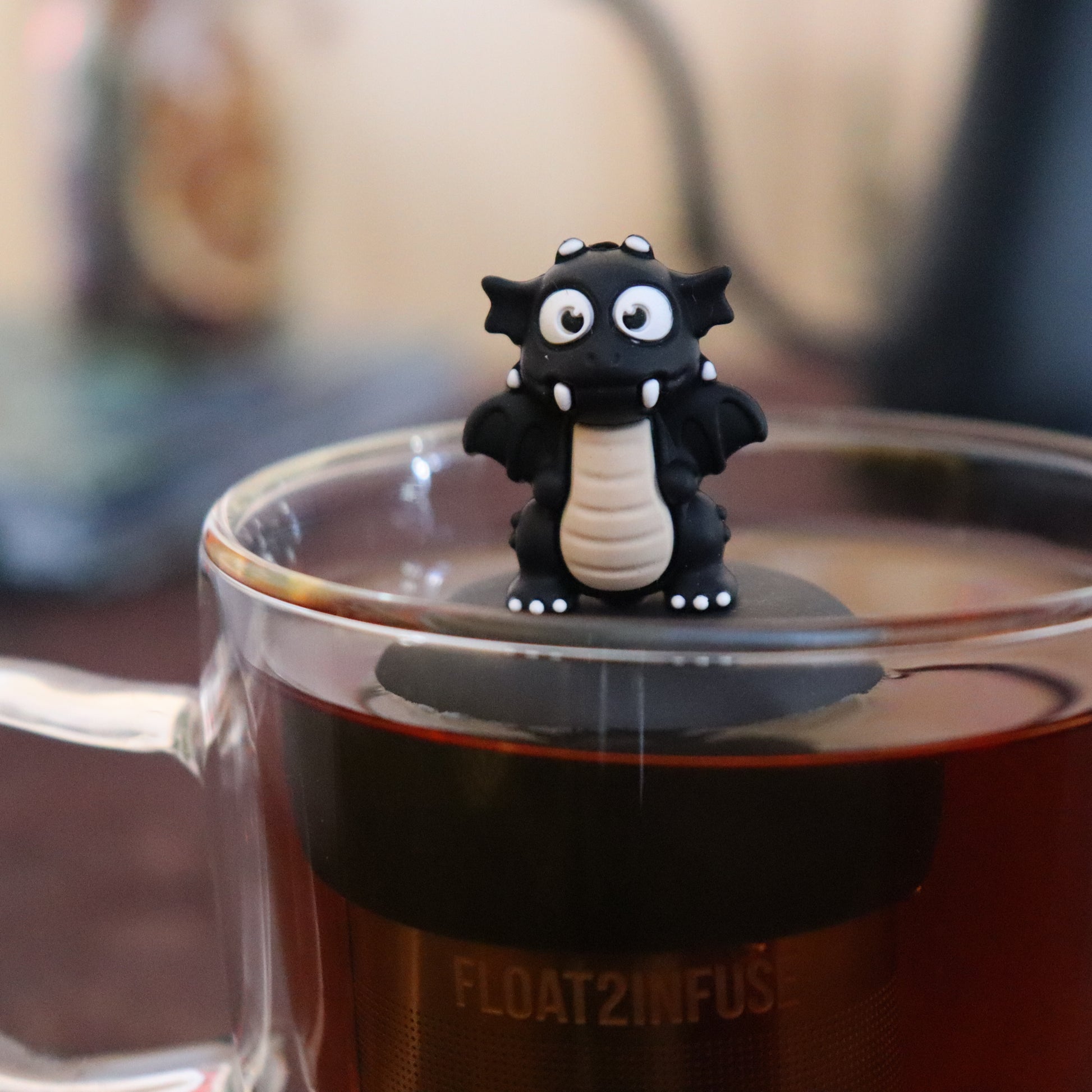 black dragon animal tea infuser for loose leaf tea  with a fine mesh stainless steel tea infuser that reads FLOAT2INFUSE on a black base floating in a glass tea mug with tea kettle in the background