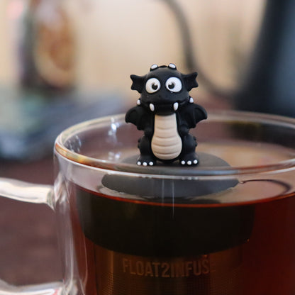 black dragon animal tea infuser for loose leaf tea  with a fine mesh stainless steel tea infuser that reads FLOAT2INFUSE on a black base floating in a glass tea mug with tea kettle in the background