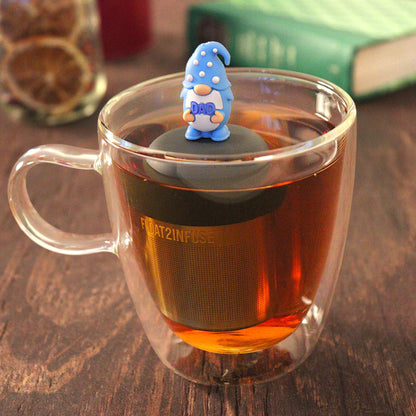 gnome with long white beard wearing blue with white polka dots holds text "dad" tea infuser for loose leaf tea with a fine mesh stainless steel tea infuser that reads FLOAT2INFUSE on a black base  floating in a glass tea mug with  loose leaf tea and books in the background